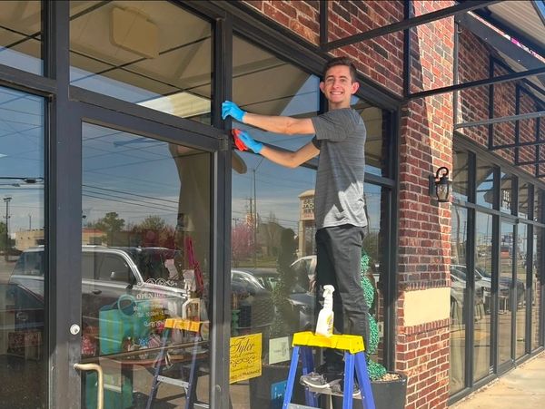 A man cleaning a storefront window while standing on a ladder outside a brick building.