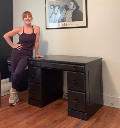 A woman smiling and leaning on a black desk in a room with wooden floors.