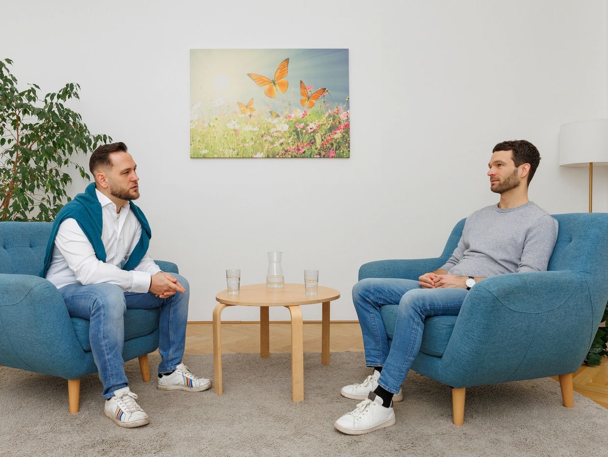 Two men sitting on blue armchairs facing each other in a bright room.