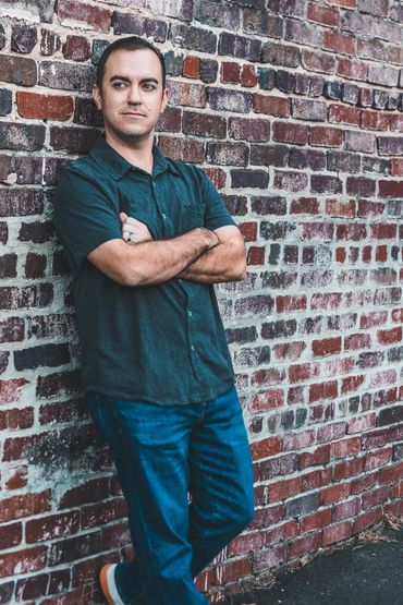 Man in casual clothes leaning against a brick wall with arms crossed.