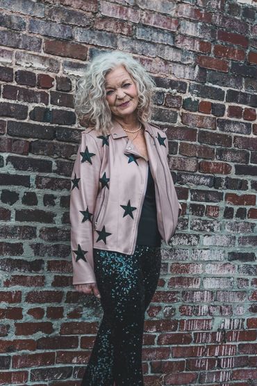 Older woman with curly gray hair wearing a star-patterned jacket poses against a brick wall.