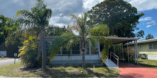 Small house with palm trees and a red driveway under a cloudy sky.