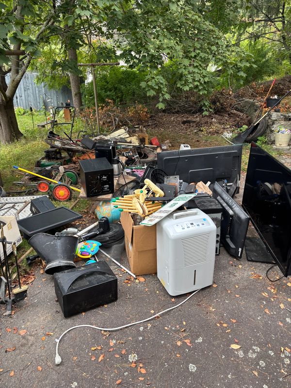 A pile of discarded electronics and household items in a driveway under trees.