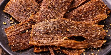 Spicy beef jerky sprinkled with crushed pepper on a dark plate.