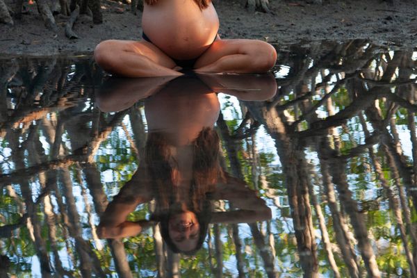 Pregnant woman sitting cross-legged near water with her reflection visible.