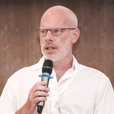 Bald man with glasses speaking into a microphone.
