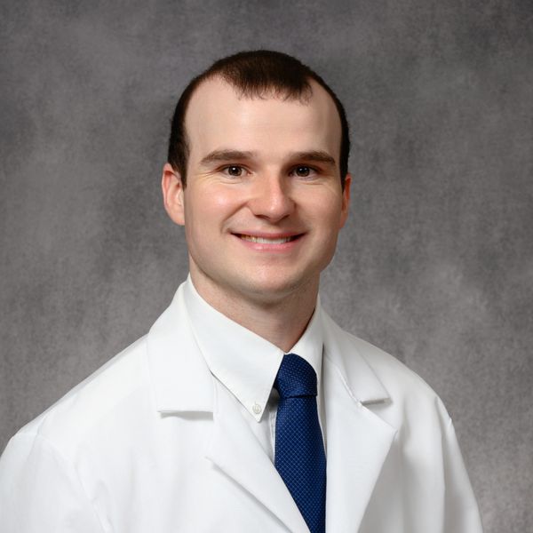 Smiling man in a white lab coat and blue tie against a gray background.