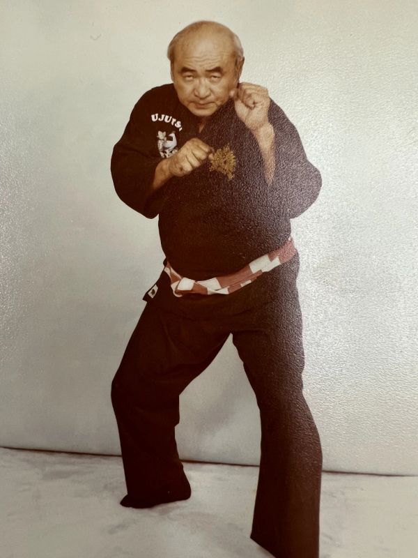 Elderly martial artist in black uniform with red and white belt in fighting stance.