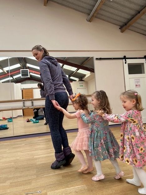 A woman leads three young girls holding hands in a dance studio.