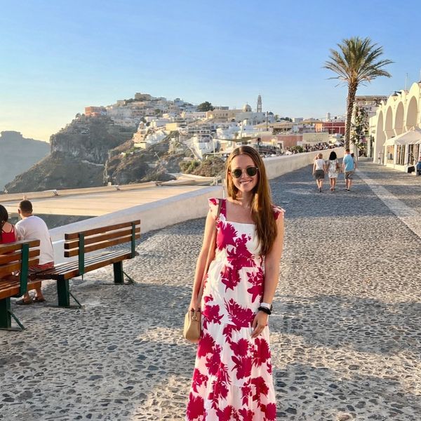 Woman in a floral dress standing on a cobblestone path with a scenic hillside town behind her.
