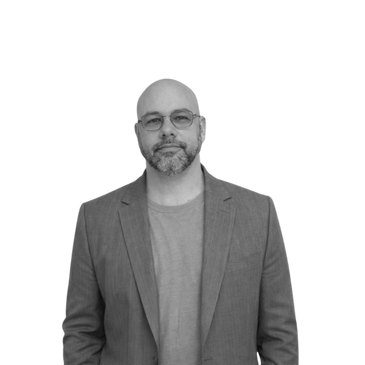 Man with glasses wearing a blazer and t-shirt, standing calmly.