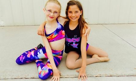 Two young girls in colorful dance outfits sitting together on concrete.