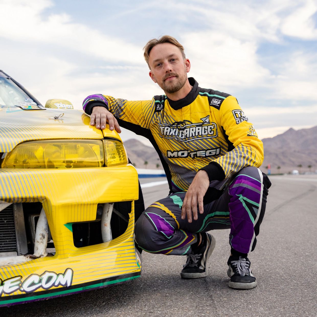 A man in a colorful racing suit kneels beside a yellow race car on a track.
