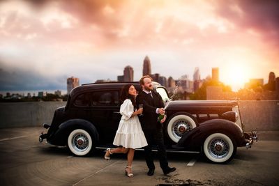 Couple dressed vintage posing beside a classic car at sunset with city skyline.