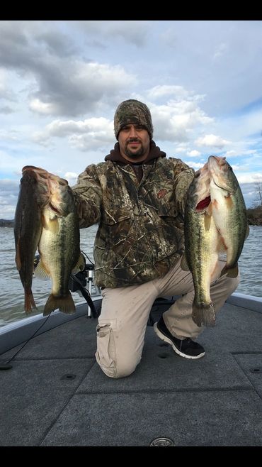 Man kneeling on a boat holding four large fish, wearing camouflage jacket and hat.