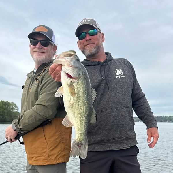 Two men proudly hold a large fish by the lake on a cloudy day.