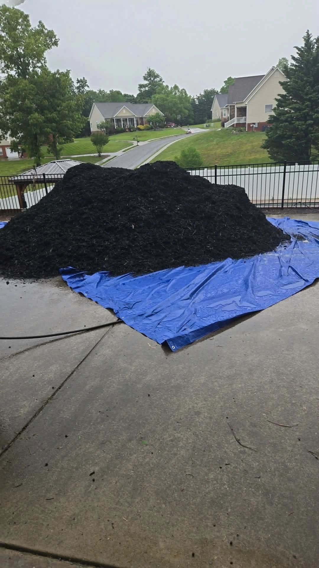 Large pile of black mulch on a blue tarp in a residential driveway.