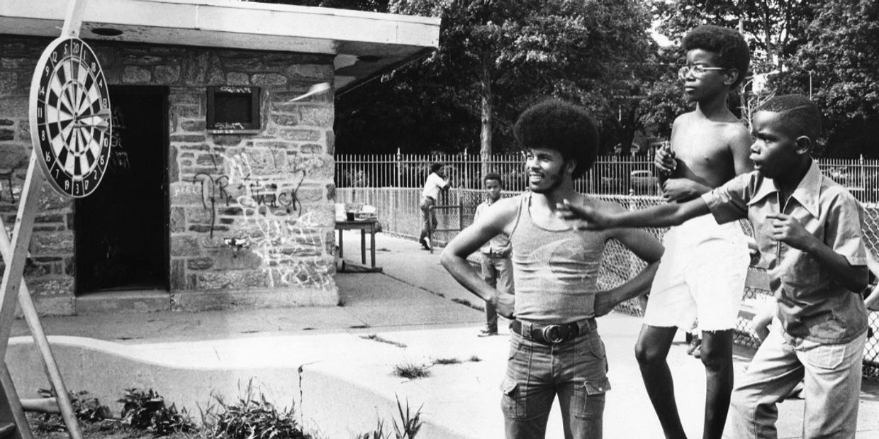 Three boys playing darts outside a stone building with graffiti.