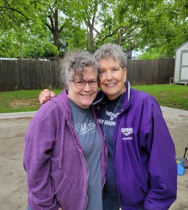 Two smiling women in purple jackets standing outdoors with trees in the background.