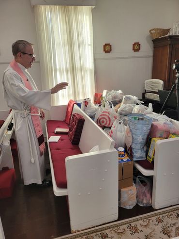 A priest in white robes blessing a pile of donated goods in a room.