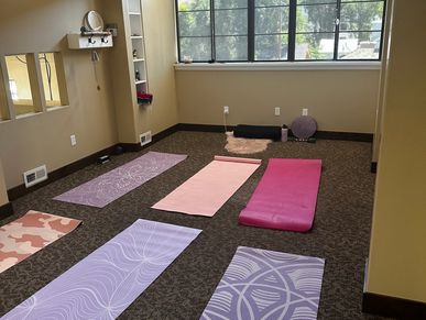 A serene yoga room with six colorful mats laid out on carpeted floor under large windows.