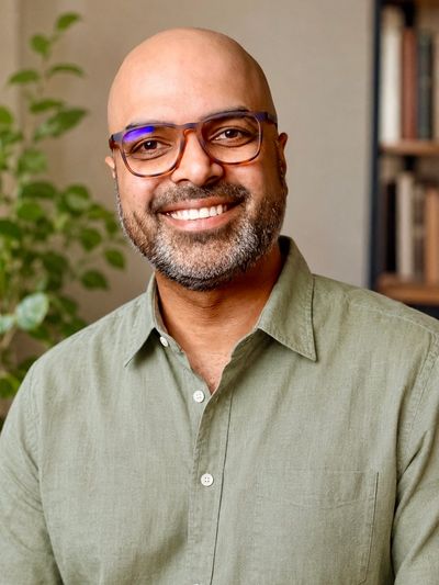 Neel Panchmatia smiling in a sage green shirt near books and plants.
