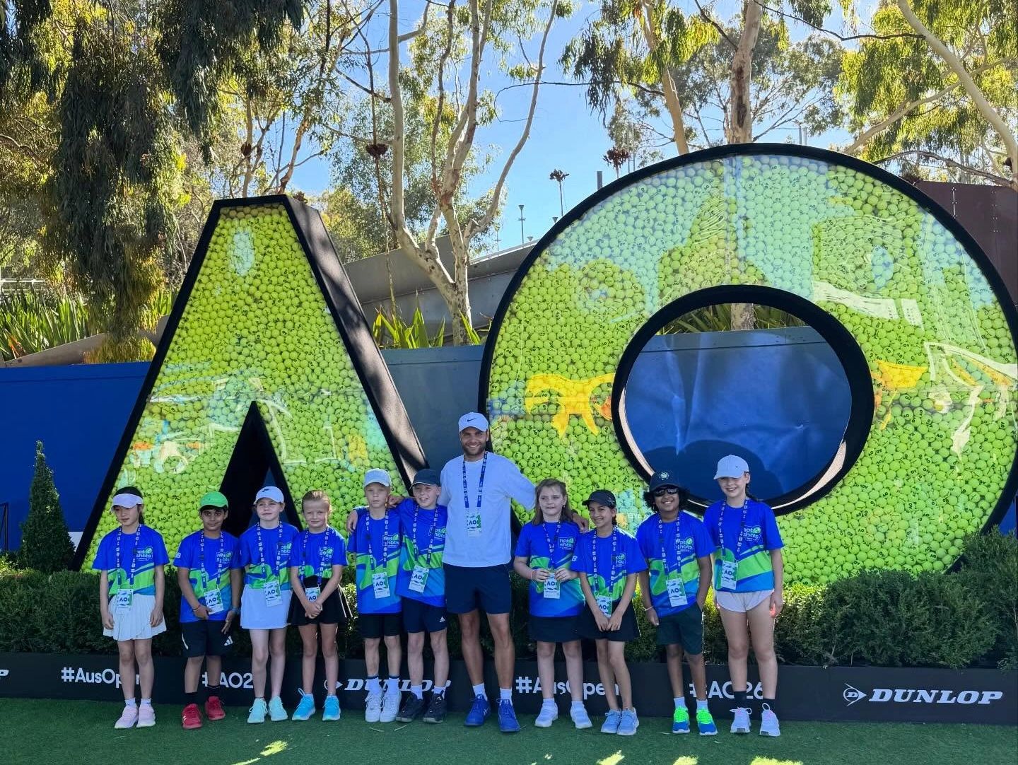 Group of kids and a coach posing by large AO tennis ball displays.