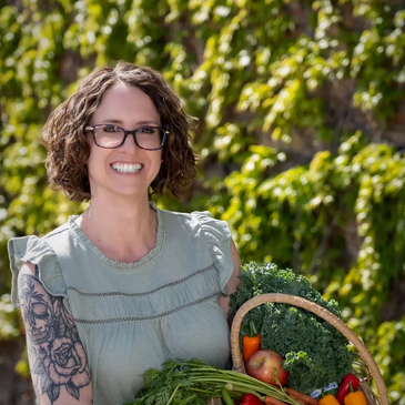 Smiling woman with glasses holding a basket of fresh vegetables outdoors.