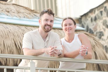 Smiling man and woman leaning on a railing outdoors with a thatched roof background.
