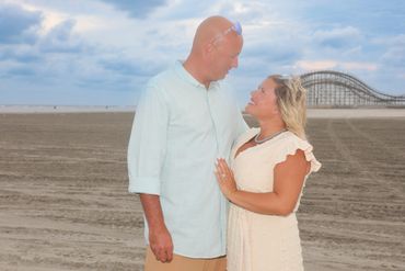 A couple gazes lovingly at each other on a beach with a roller coaster in the background.