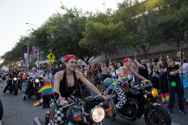 Two women ride motorcycles adorned with rainbow flags in a lively pride parade.
