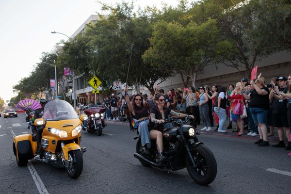 Two women ride a motorcycle during a lively parade with cheering spectators.