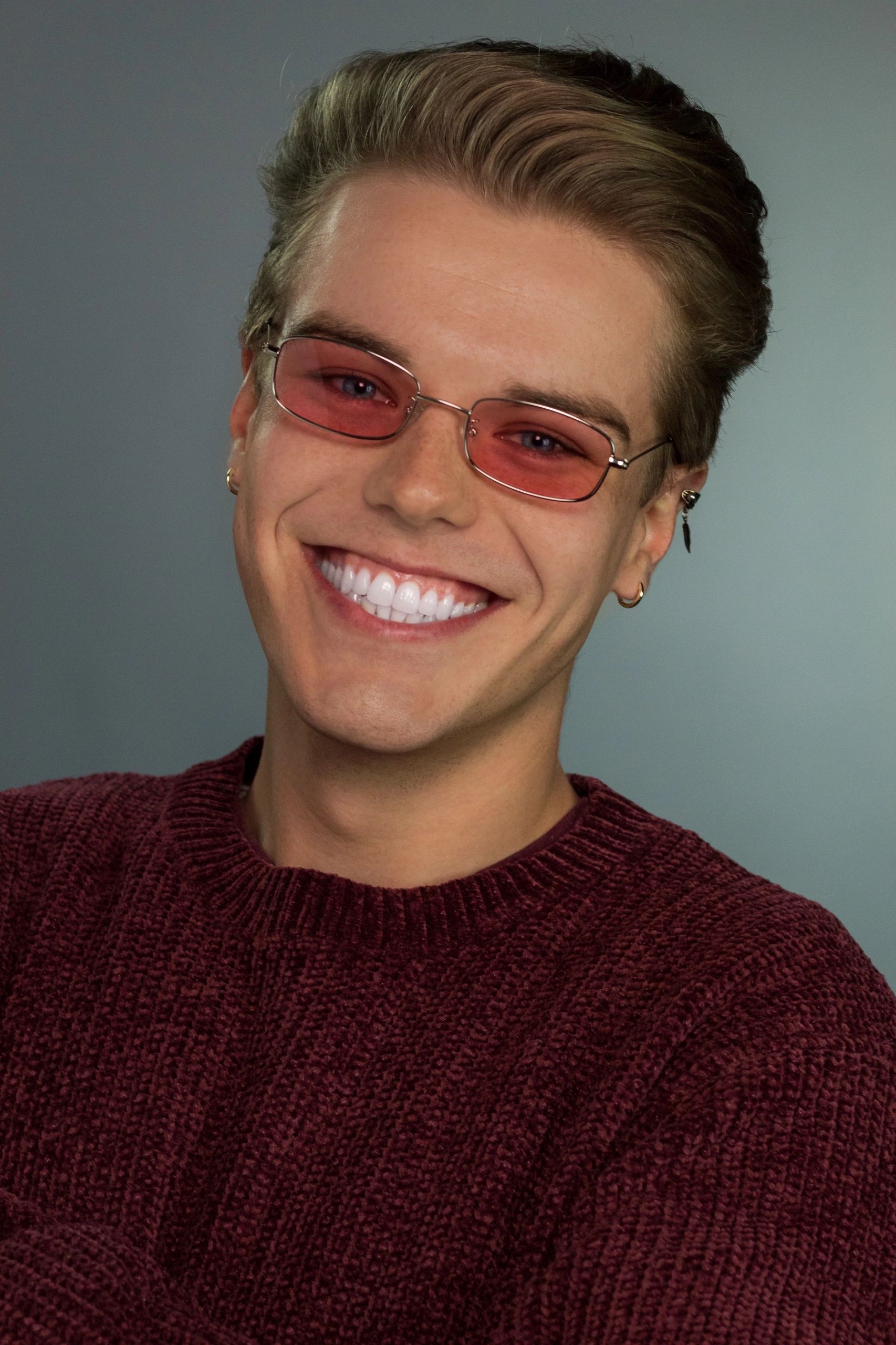 Smiling young man wearing pink-tinted glasses and a maroon sweater.
