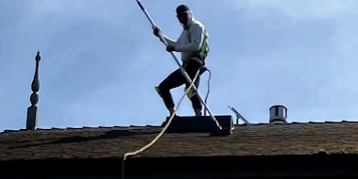 Worker on a rooftop using a long tool under clear blue sky.