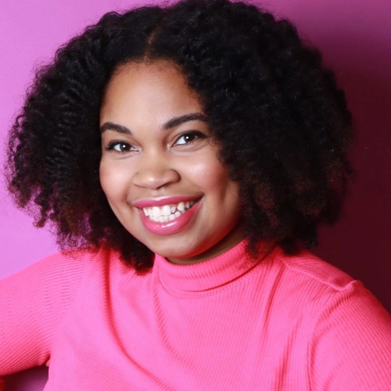 Woman with curly hair smiling in a pink top against a pink background.
