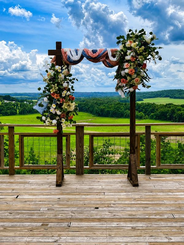 Floral wedding arch on a wooden deck with scenic green fields and blue sky.