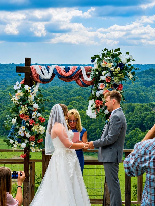 Couple exchanging vows under a floral-decorated arch with a scenic mountain backdrop.