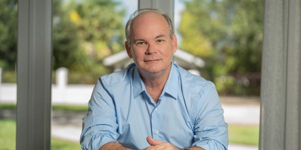 A man in a light blue shirt sitting indoors with a garden visible outside.