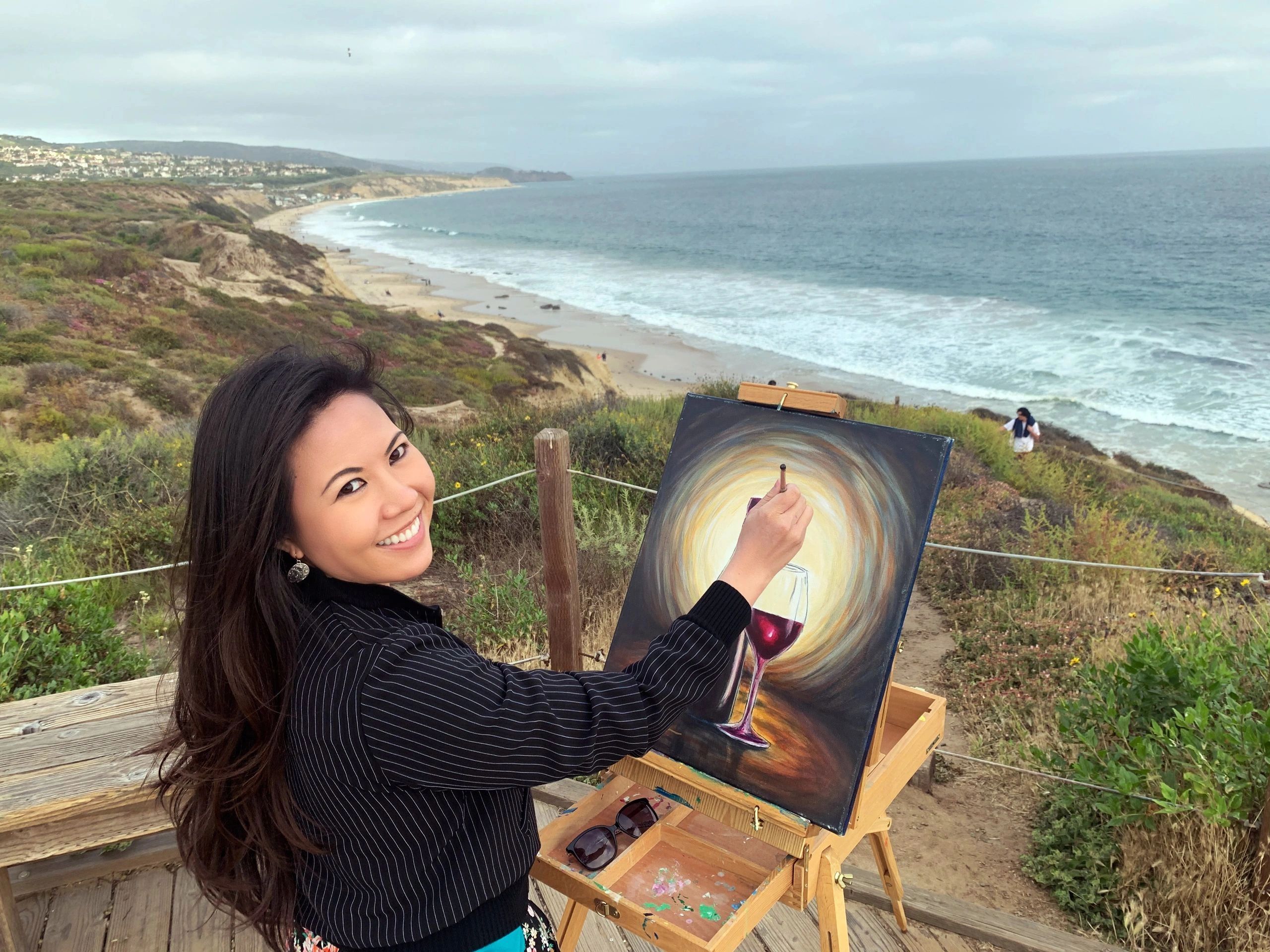 A woman paints a wine glass on a canvas by the beach with a smile.