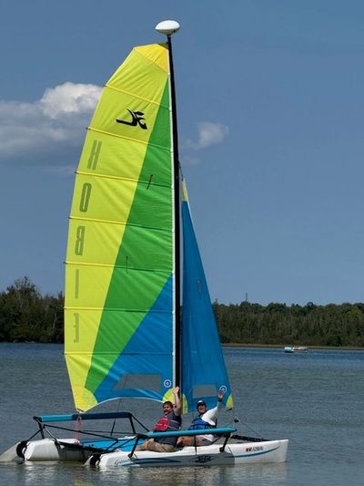 Two people sailing a colorful Hobie catamaran on calm water.