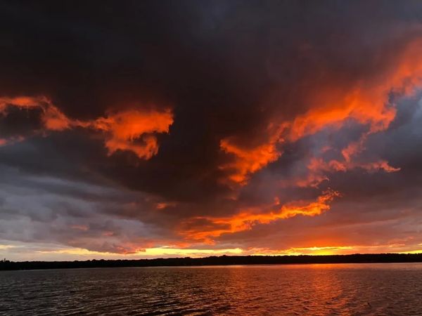 Fiery orange and red sunset reflecting on a calm lake under dark clouds.