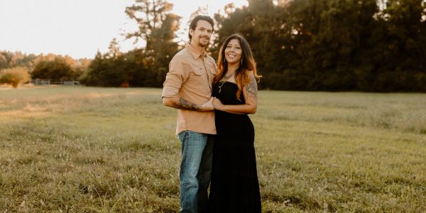 Couple standing in a grassy field during golden hour, smiling warmly.