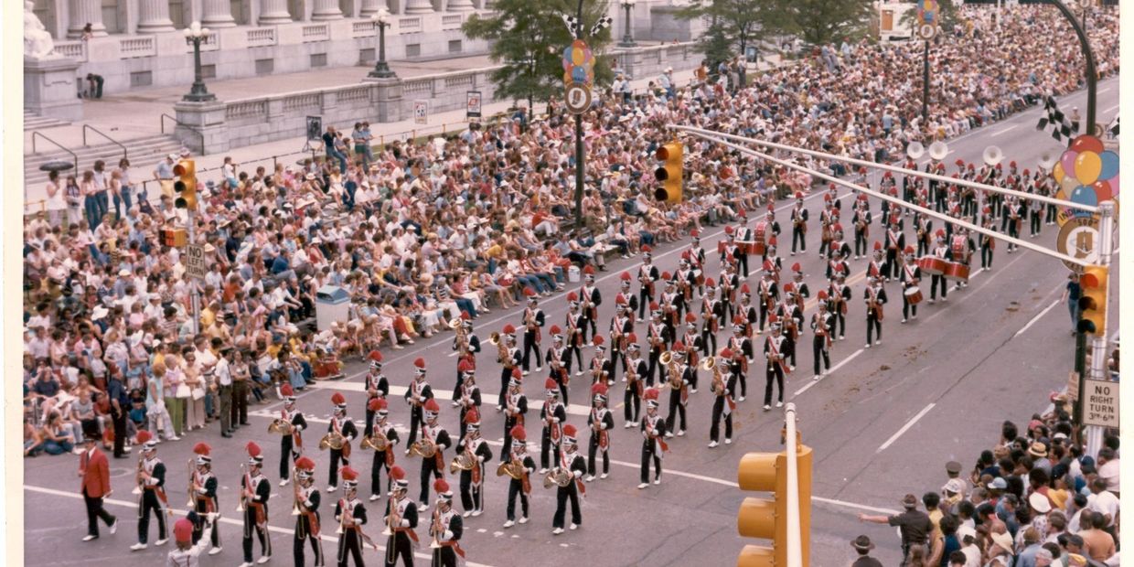 Marching band parades down a city street with large crowd watching.