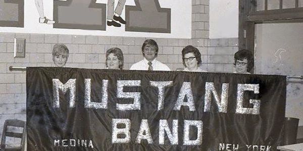 Five people behind a banner that reads 'Mustang Band, Medina, New York'.