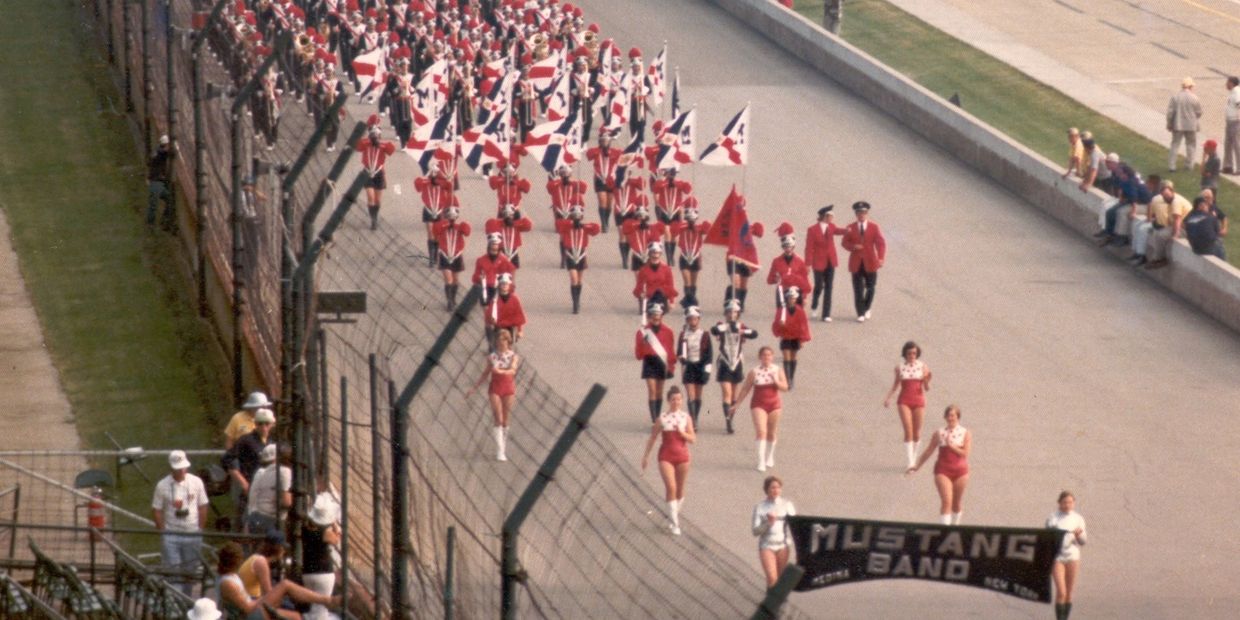 Mustang Band marching in formation with flags and uniforms on a raceway.