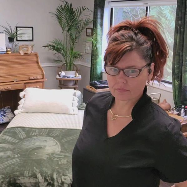 Woman in glasses standing in a cozy room with plants and a bed.