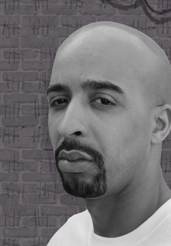 Close-up of a serious bald man with a goatee against a brick wall marked with tally marks.