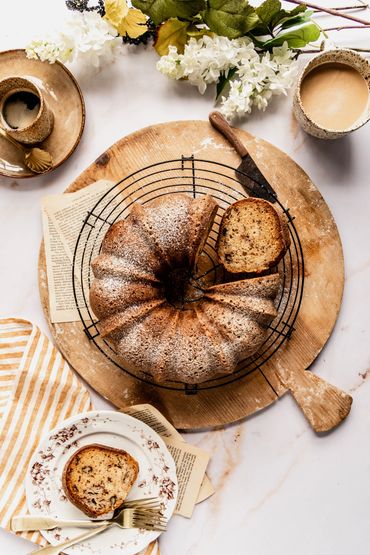 Bundt cake with a slice cut, coffee, and flowers on a wooden board.