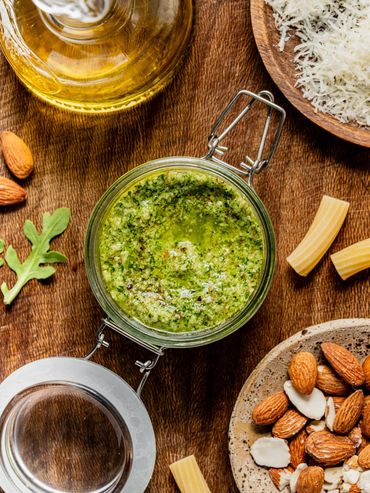 Jar of green pesto sauce surrounded by ingredients on wooden table.