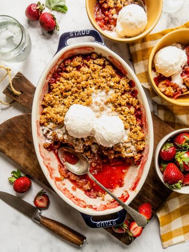 A fruit crumble dessert topped with scoops of vanilla ice cream and fresh strawberries on the side.